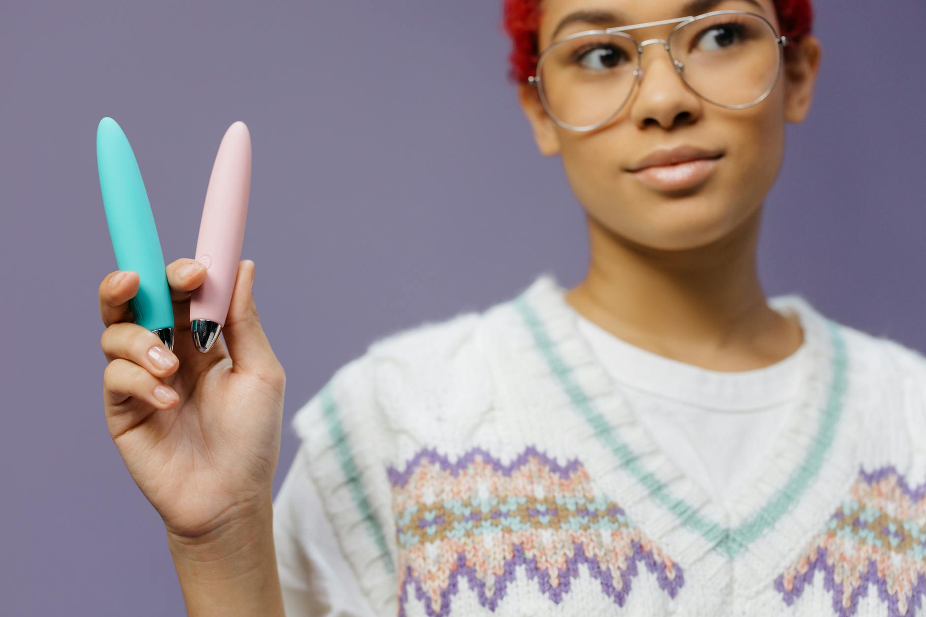 A young woman with eyeglasses holds two colorful vibrators against a purple background.